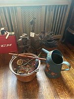 Photo showing a painted metal watering can with floral decoration, two metal sculptures (rooster and man), a copper bucket with small items, and surrounding wooden floor and furniture.