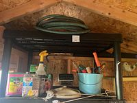 Photo of garden supplies on black plastic shelving including garden hose, plant food containers, garden tools in blue bucket, and garden figures.