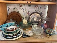 Kitchen shelf displaying an assortment of glass bowls, ceramic plates with bird designs, a large silver serving tray, and a covered sugar bowl.
