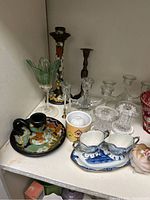 Photo of shelf with decorative candle sticks, ceramic mugs, and porcelain ramekins including a floral cream and sugar bowl set and a unique black and yellow candle holder.