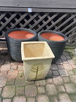 Three plastic garden planters arranged outdoors on a stone patio in front of lattice fencing: two large round black textured planters with orange interiors, and one smaller square beige planter with tree design.