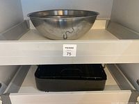 Photo shows largest stainless steel mixing bowl on shelf above black rectangular roasting pan with handles.