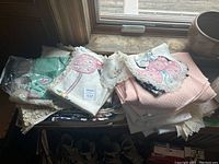 Stack of various folded tablecloths and some napkins piled on a small surface by a window. Includes embroidered, lace trimmed, and pink textured fabrics.