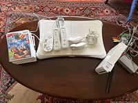 Overall view of Wii Fit Balance Board with two Wii remotes, two Nunchuks, classic controller, game cases, and cables on a wooden table.