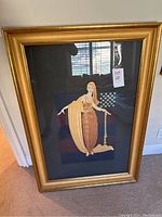 Front view of framed print leaning against wall, showing gold-tone frame, black mat, Art Deco female figure