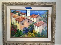 Full view of the framed painting showing houses with red roofs and surrounding colorful plants, with framed and a linen liner.