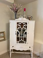 Front view of white antique hutch cabinet showing decorative glass door, single drawer, and legs with crossbar.