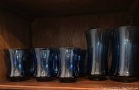 Photo showing multiple blue glass drinking vessels of varying heights and shapes arranged inside a wooden cabinet shelf.