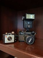 Both cameras displayed on a wooden shelf, showing front and side details of Ricoh Auto TLS EE camera with top flash and Kodak Brownie Holiday Flash camera next to it.
