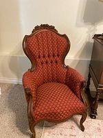 Front view of an antique Victorian armchair with red patterned upholstery and carved mahogany wood frame, showing button tufting and floral crest.
