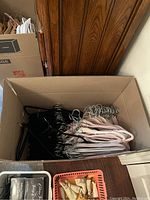 Open cardboard box filled with multiple felt hangers in black, cream, and rose colors showing metal hooks stacked inside.