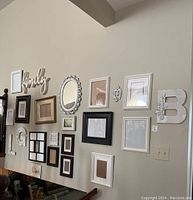 Gallery wall with assorted empty picture frames of various sizes, colors, and shapes including white ornate oval and rectangular frames and decorative letters 'family', 'G', and 'B'.