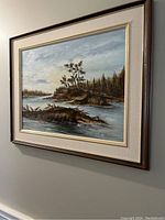 Framed landscape painting with river, trees, and rocky banks shown hanging on wall, framed in brown wood with beige mat and cream trim.