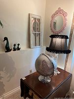 Three Blue Mountain duck figurines on ledge with distressed white window frame mirror and pink ornate mirror on wall, a silver globe and lamp with black shade on table