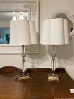 Set of two lamps on a wooden surface under decorative mirrors, showing glass and brass bases with cream scallop fabric shades.
