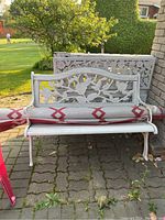 Outdoor white painted metal and wood bench with floral design backrest and curved arms with rolled red and white patterned outdoor carpet on seat.