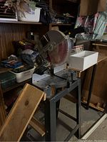 Mitre saw mounted on a grey metal stand with a wood plank leaning nearby, showing the saw blade and garage storage environment.