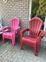 Two Adirondack-style plastic chairs, one pink and one red, showing dirt and outdoor wear on a paved surface near a brick wall.