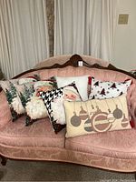 Six Christmas themed throw pillows with Santa Claus faces, trees, ornaments, and holiday patterns arranged on pink couch.