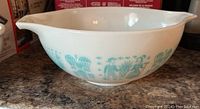 Side view of off-white Pyrex mixing bowl with teal Amish Butterprint pattern showing vintage people and foliage designs.