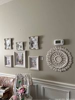 Six white ornate picture frames and large white carved wall medallion displayed on wall with digital thermostat above medallion and chair with floral upholstery below frames.