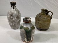 Three decorative art pottery pieces together: a tall gray patterned vase, a small green and white jar with lid, and a medium brown handled jug.