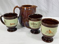Set of three brown and cream-colored ceramic coffee mugs with raised hunting scenes and a matching brown pitcher shown from front