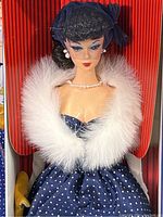 Front close-up showing Barbie doll with navy polka dot dress, white fur stole, pearl jewelry, and veiled hat, standing in striped red display box.
