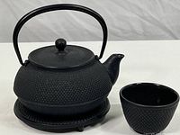 Angle showing the black cast iron tea pot on its matching trivet with the lid on, view of the textured exterior and curved handle.