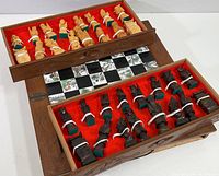 Open wooden carrying case with red felt-lined drawers holding light and dark carved chess pieces and partially visible board.