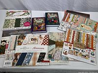 Large assortment of several scrapbook paper pads featuring different seasonal and thematic designs, and multiple sticker sheets laid out side by side on a white cloth.