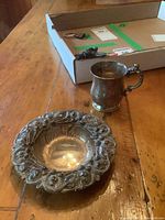 Both the antique sterling silver baby mug and the ornate nut bowl on a wooden table.