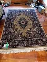 Full view of rectangular area rug showing overall pattern and fringed ends, placed on wooden floor with some surrounding household items.