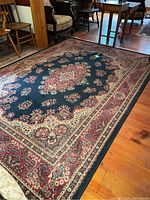 Full view of a 10x6 feet rectangular area rug with floral pattern, dark blue central field and red border, placed on hardwood floor near chairs.