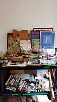 Overall view of two-tier art supply cart and adjoining surface with palettes, brushes, paints, sketchbooks and tools