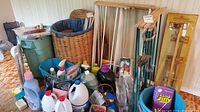 Wide view of cleaning products including spray bottles, bleach, detergent, and other bottles. Large wicker laundry hamper with blue fabric lining is visible, along with wooden drying racks and several other household items.