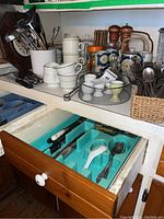 Photo of counter showing various kitchen items including cooking utensils in white ceramic holder, teapots with matching cups, metal basket with egg cups, salt and pepper shakers, and open drawer with cutlery.