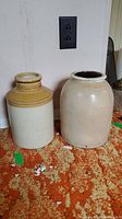 Photo showing two earthenware jugs side by side on orange patterned carpet against white wall with electrical outlet visible.
