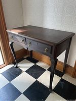 Front angle view of vintage wooden hall table showing two drawers with brass pulls and curved legs, sitting on black and white checkered floor with patterned wallpaper background.