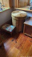 Wicker hamper and upholstered foot stool shown on wooden floor next to wooden furniture.