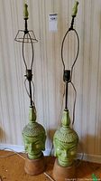 Pair of green ceramic Buddha head lamps on wooden bases with metal lamp harps and no shades, standing against a striped wallpaper.