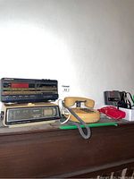 SoundDesign cassette clock radio, Sanyo 2 band clock radio, beige rotary phone, red rotary phone, and Sony Walkman with headphones on a wooden surface.
