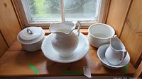 Photo of the complete set on a wooden window ledge showing all five pieces: large wash basin with pitcher, covered pot, small pitcher on bowl, and an extra bowl.