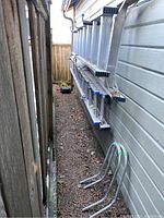 Photo showing two aluminum ladders leaning vertically against an exterior wall, one shorter (approx. 8 ft) and one longer (approx. 12 ft). Both ladders show dirt and outdoor wear.