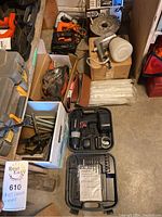 Overall view of belt sander, power tools, sanding belts, drill in black case, and other boxes with tools and wiring items in garage setting.