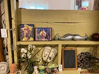 Wide shelf shot with various decor items including two surreal art prints, a silver mustache clock missing top half, small chalkboard and figurines