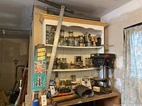 Full view of wooden shelving unit and workbench with TMT drill press, storage jars with nails and screws, and various hand tools, including a metal ruler and a box of nails.