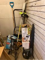 Photo shows the collection of garden tools in corner including sprayer box, leaf blower, shovel with turquoise handle, small metal wheeled cart, manual mower, and garden bags.