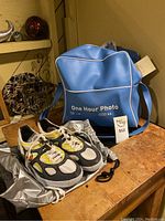 Pair of Nike sneakers placed on gray fabric bag next to a blue "One Hour Photo" shoulder bag with white piping.