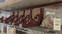 Photo showing multiple wooden shelf brackets painted reddish-brown with ornate scroll cut-outs, aligned on a rack with a plastic bag of screws.